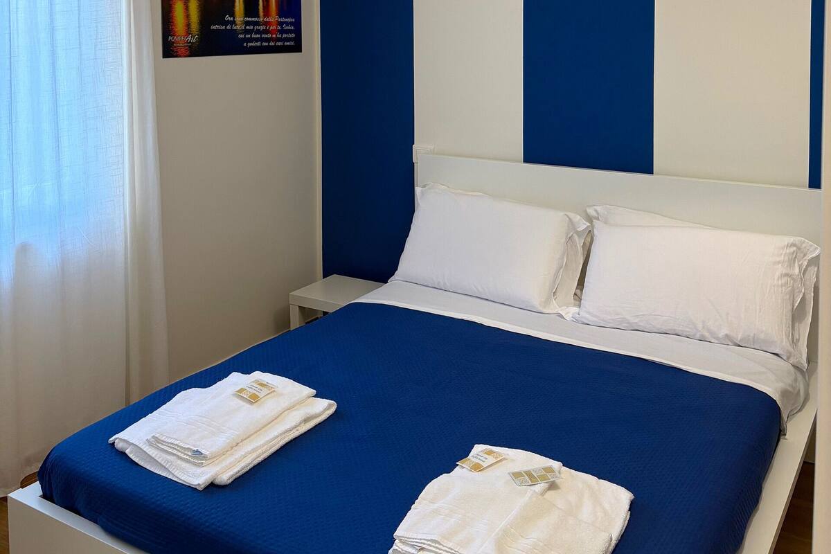 A hotel room with a bed, white pillows, a blue bedspread, and a white towel on the bed.