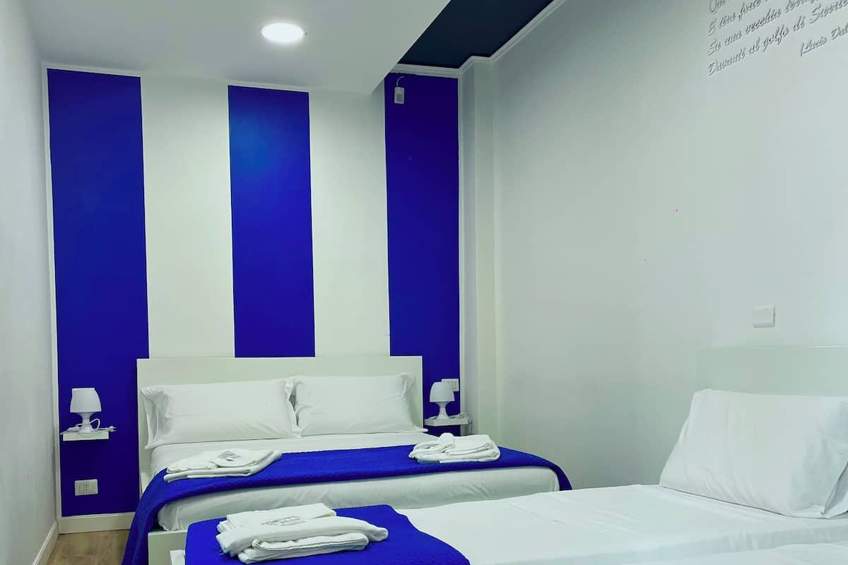 A hotel room with two beds, each with white linens and blue bedspreads. There are white towels folded on each bed. The room has a wooden floor and white walls with blue accents.