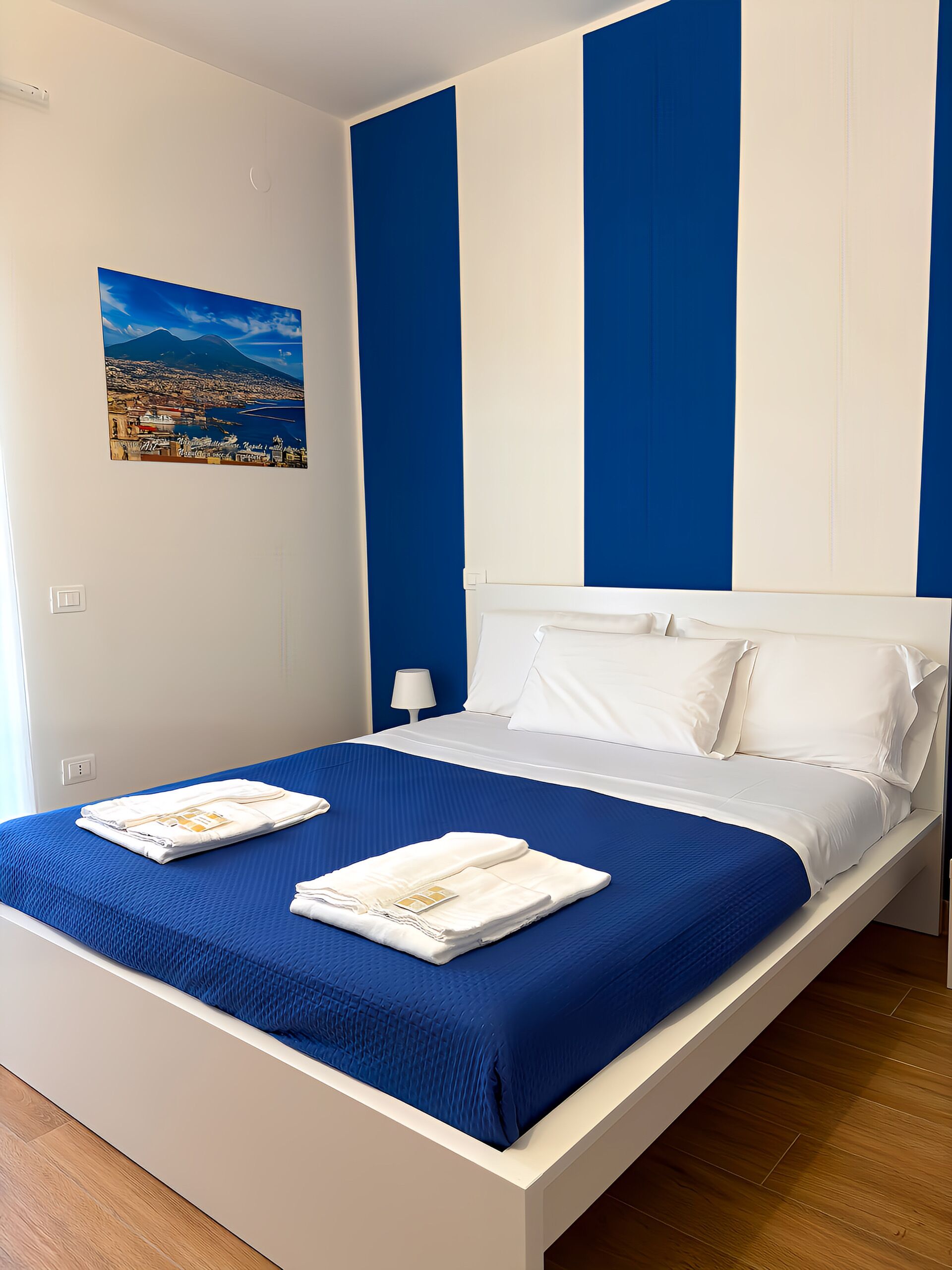 Una cama bien hecha con sábanas blancas y una colcha azul, una mesita de noche blanca con una lámpara, y un cuadro de un paisaje urbano en la pared.