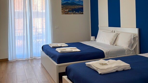 A hotel room with two beds, a large window with sheer curtains, a painting of a landscape, and a blue and white striped wall.