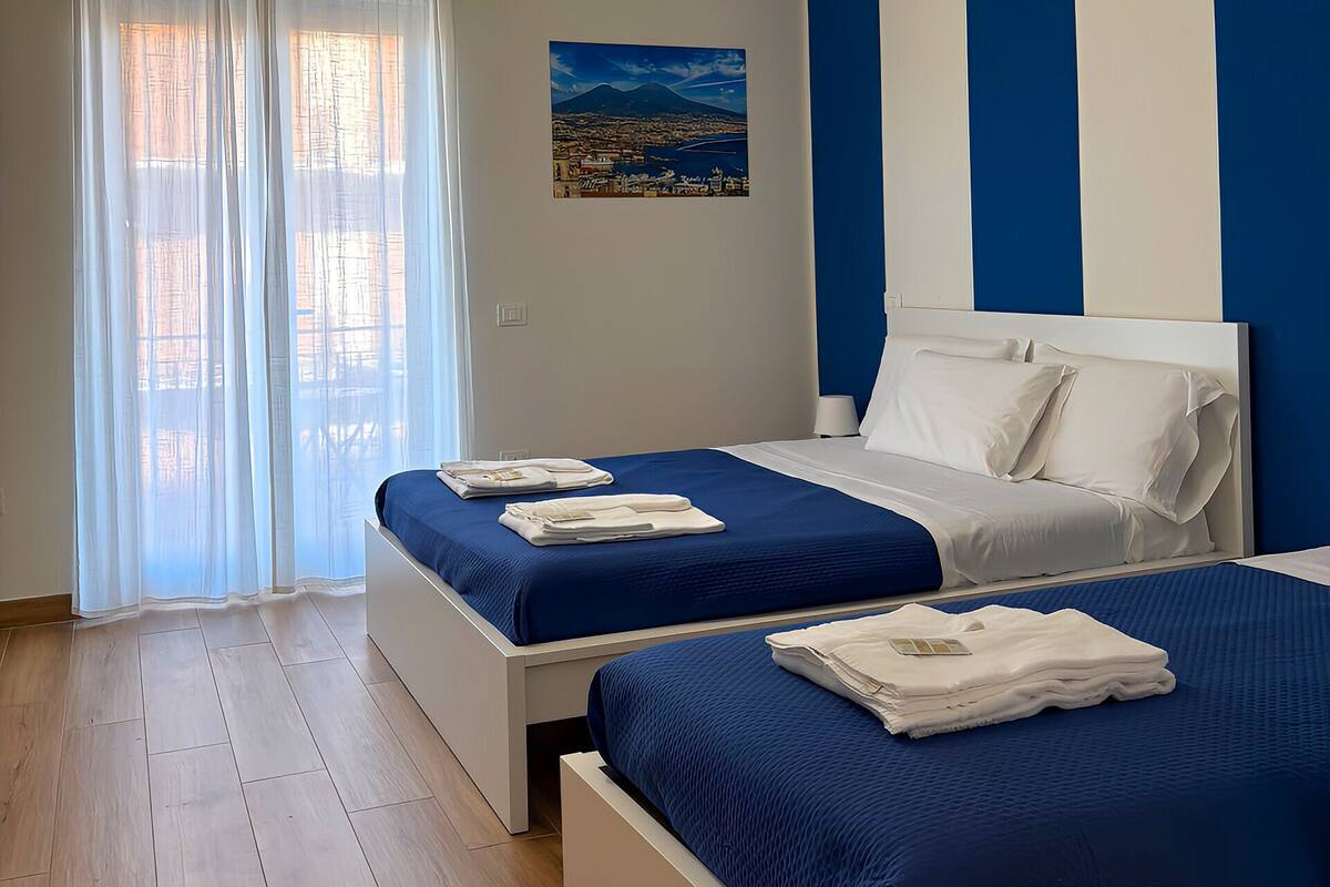 A hotel room with two beds, a large window with sheer curtains, a painting of a landscape, and a blue and white striped wall.