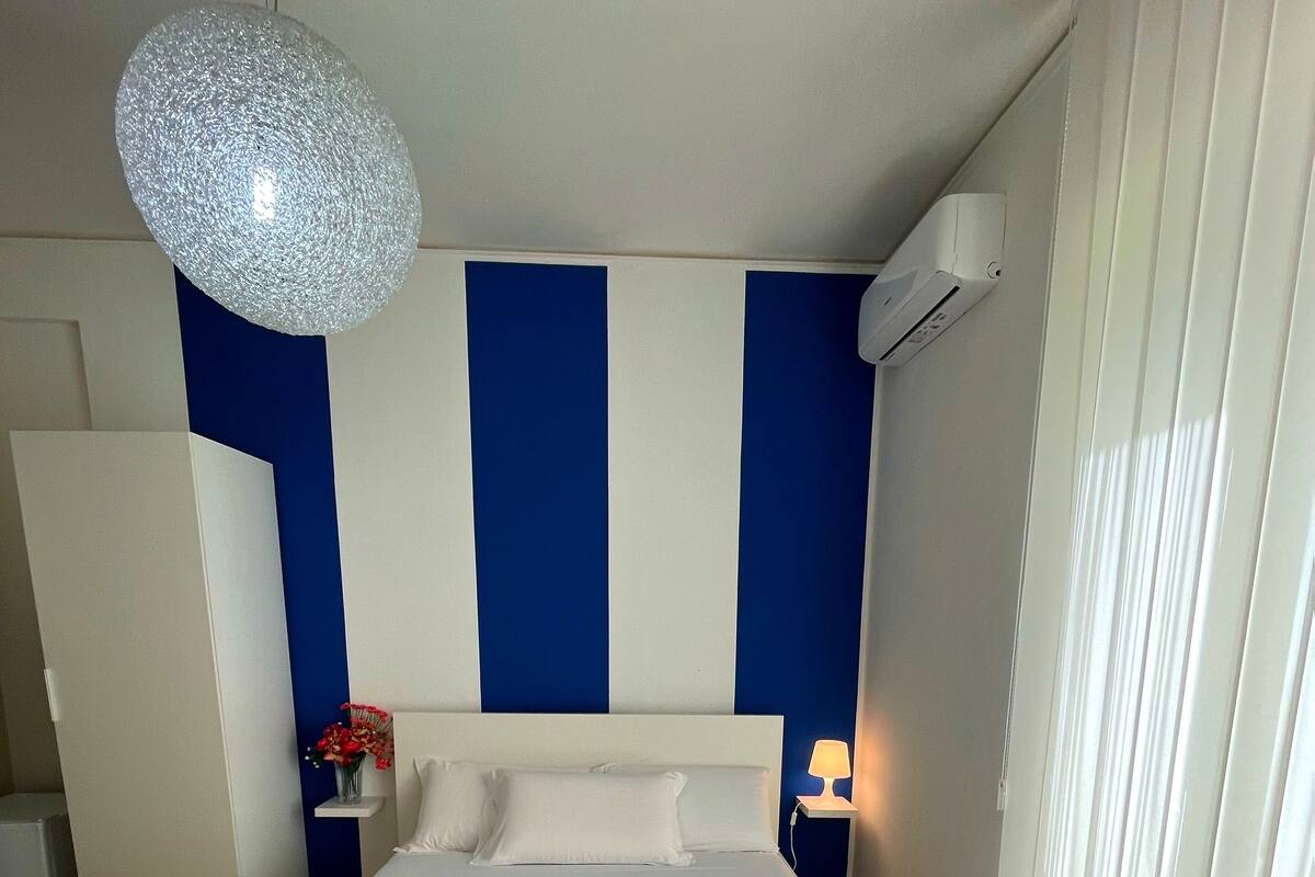 A bedroom with a large bed, a blue and white striped wall, a hanging light fixture, and a nightstand with a lamp.