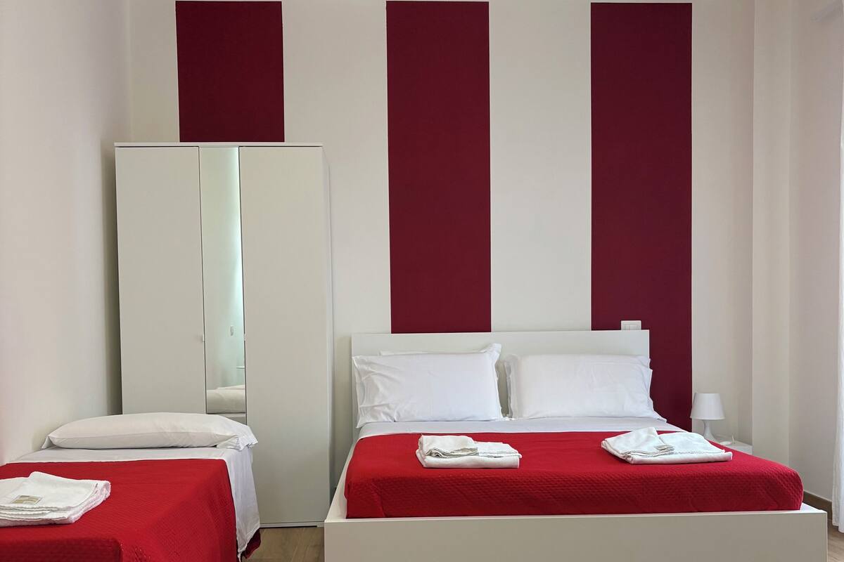 A hotel room with two beds, each with a red bedspread and white pillows. There is a large mirror on the wall, a white wardrobe, and a red accent wall.