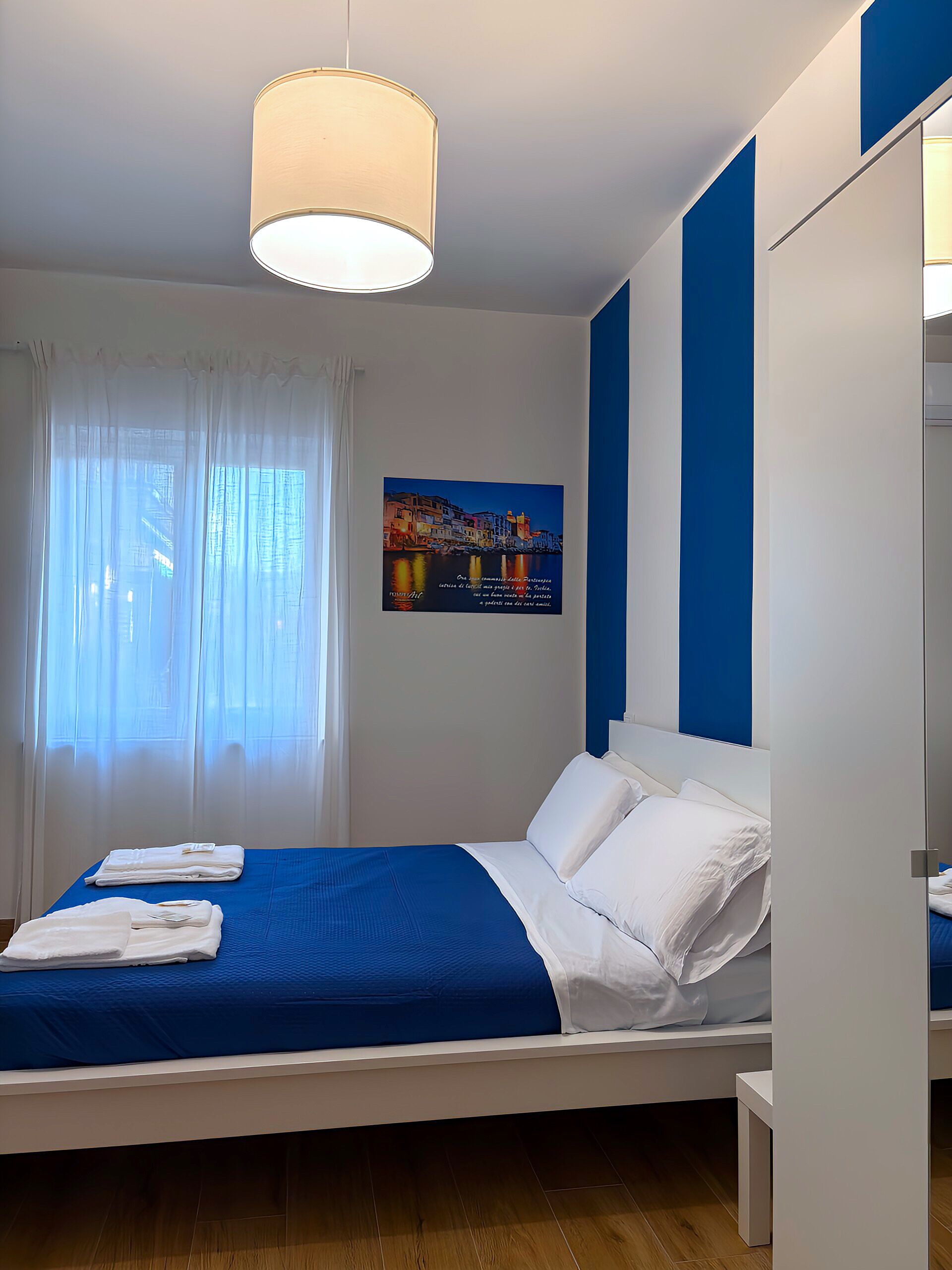 A neatly made bed with white linens and a blue bedspread, a large window with sheer curtains, and a painting of a coastal scene on the wall.