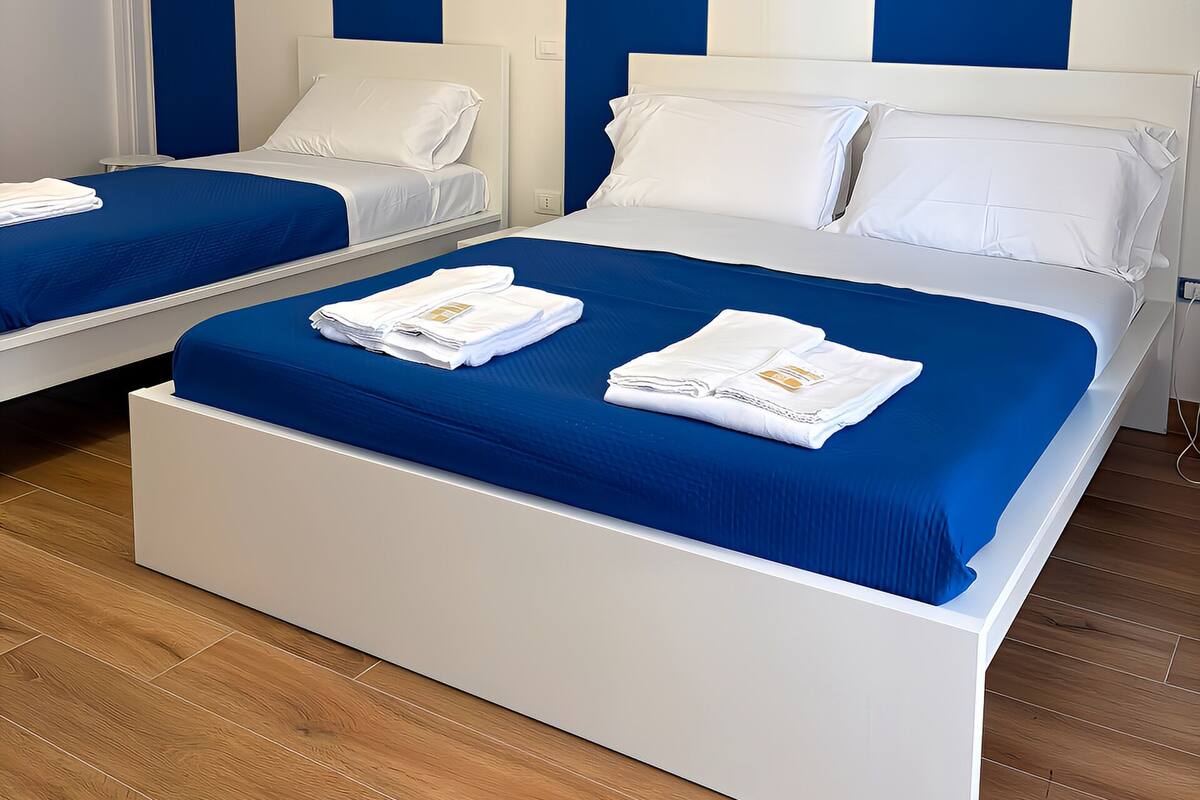 A hotel room with two beds, each with a blue bedspread and white pillows. There is a white headboard and a wooden floor.