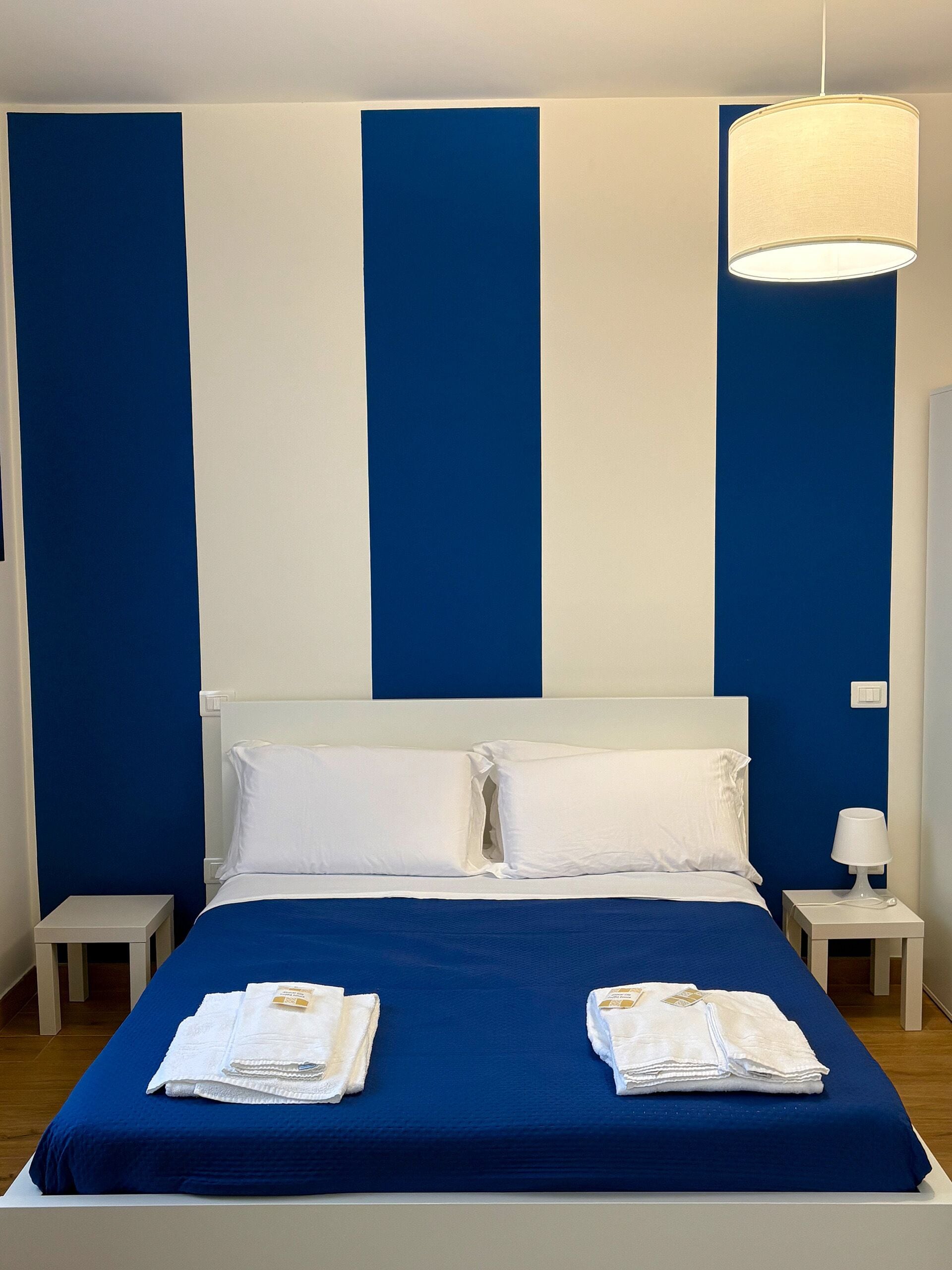 Ein Hotelzimmer mit einem großen Bett, weißen Kissen, einer blauen Bettdecke und einem weißen Nachttisch mit einer Lampe.