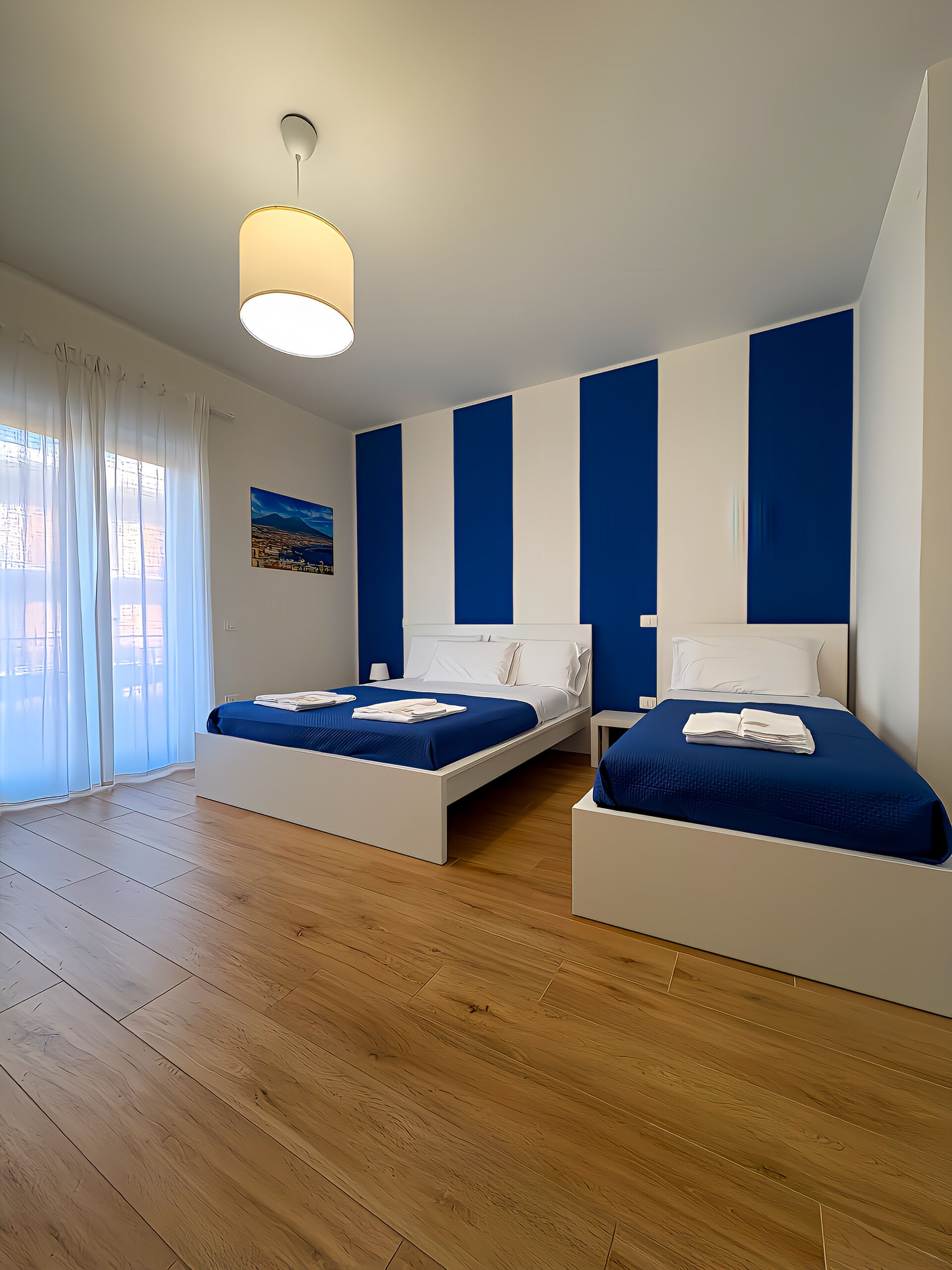 Una habitación de hotel moderna con dos camas, suelo de madera y un ventanal con cortinas translúcidas.