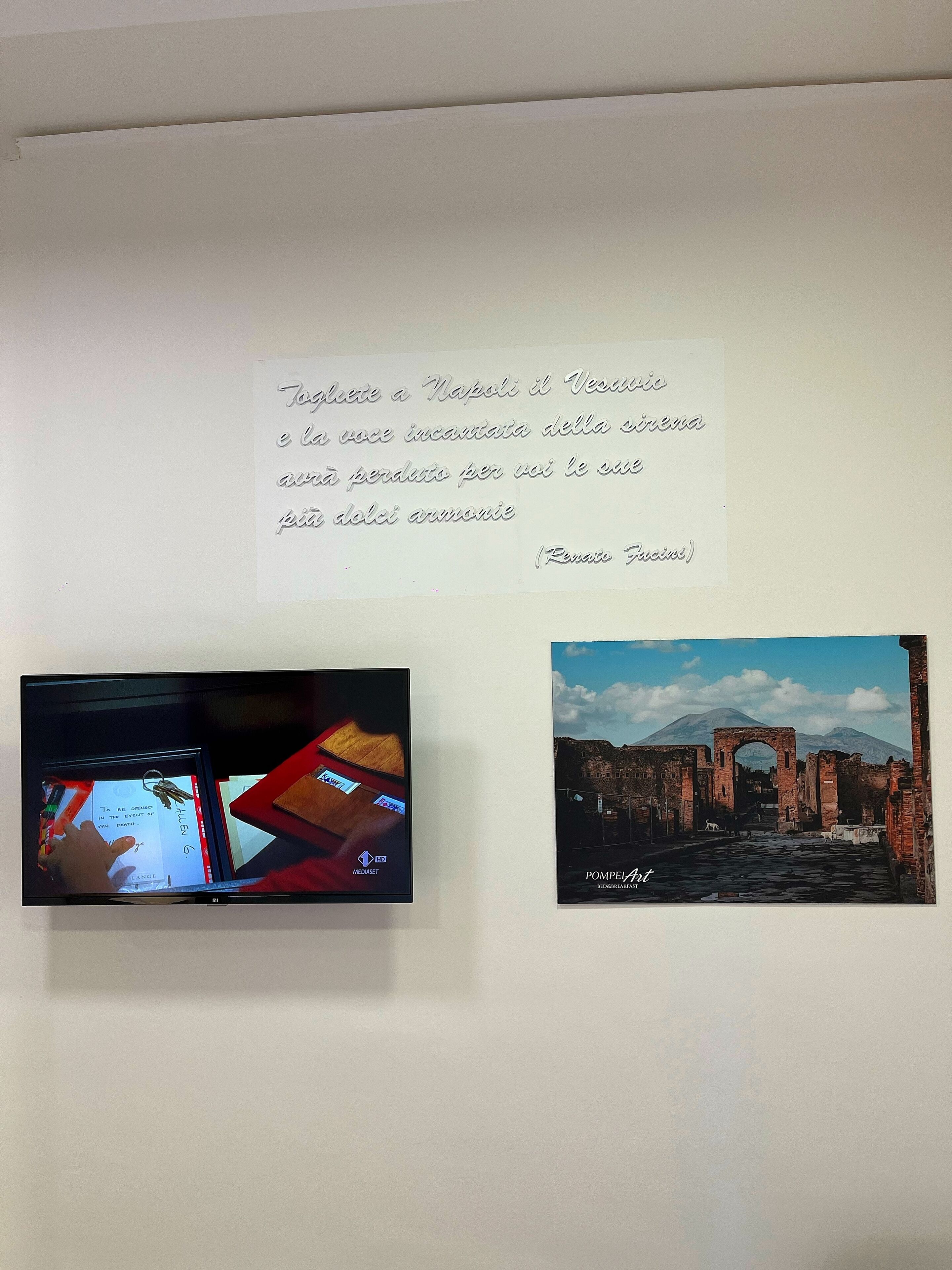 A wall with a handwritten quote in Italian, a television displaying a digital interface, and a framed photograph of a historical site.