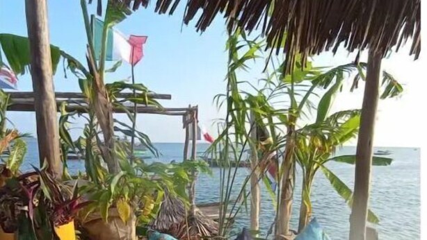 Eine Holzterrasse mit einem Reetdach, eine Sitzgelegenheit mit gemusterten Kissen und Blick aufs Meer.