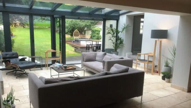Un salón amplio con un sofá grande de color gris, una mesa de centro y un comedor visible a través de un vidrio que da a un jardín.