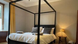 Una cama con dosel, con una colcha y almohadas blancas, una mesita de noche con una lámpara y un cubo de basura.