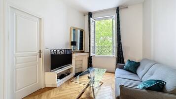 Una sala de estar con sofá, mesa de centro de vidrio, televisor y ventana con cortinas.