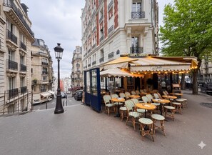 An outdoor cafe with wicker chairs and tables, a covered seating area, and classic European architecture in the background.