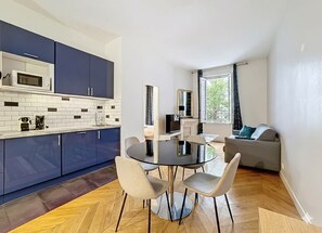 A modern kitchen with blue cabinets, a round glass table, and a grey sofa in a bright living area.