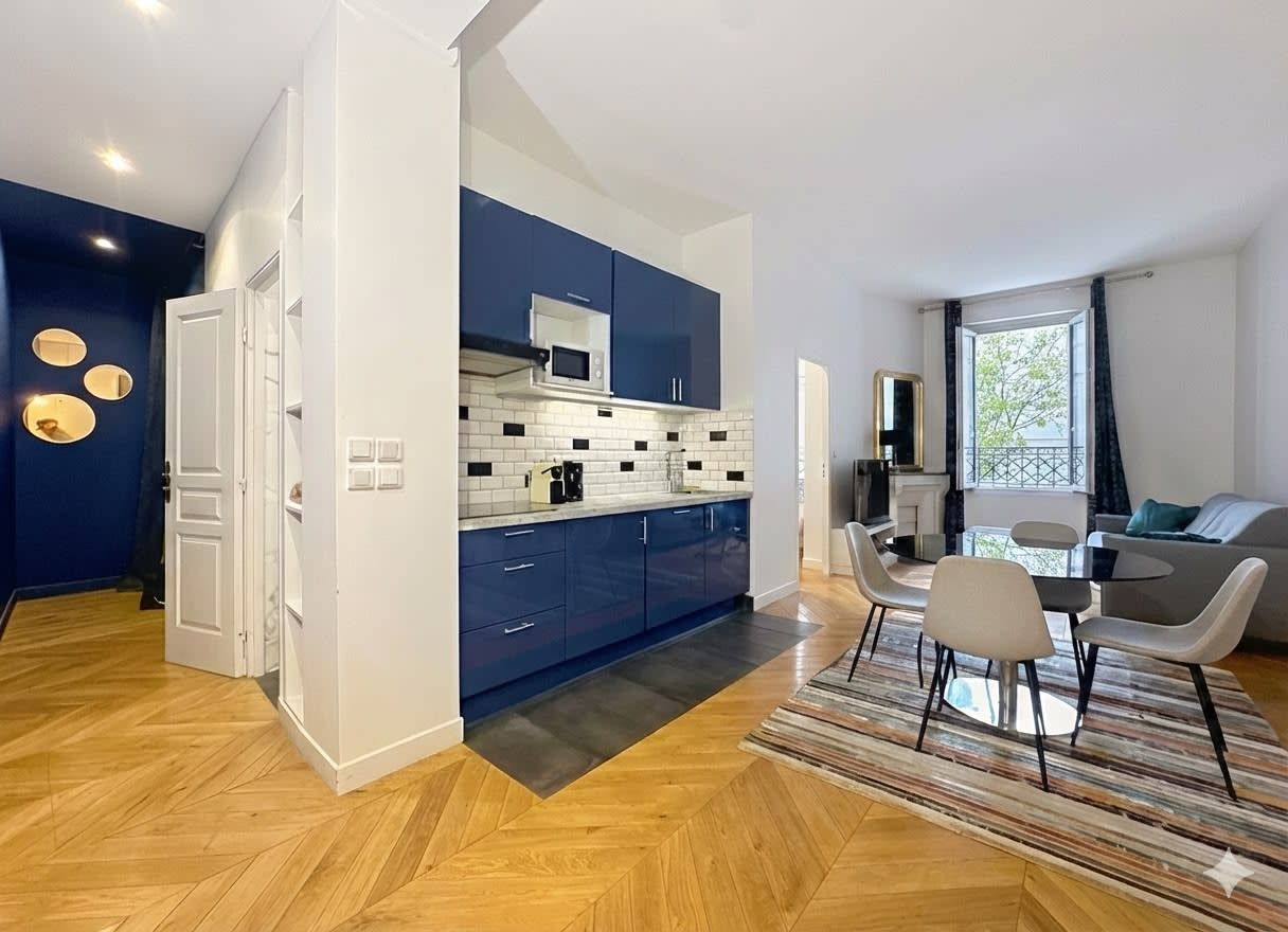 Une cuisine moderne avec des armoires bleues, un comptoir blanc et un four à micro-ondes. On y trouve un coin repas avec une table et des chaises, ainsi qu’un salon avec un canapé et une fenêtre.