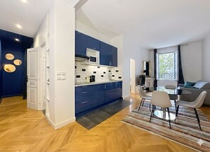 A modern kitchen with blue cabinets, a white countertop, and a microwave. There is a dining area with a table and chairs, and a living room with a sofa and a window.