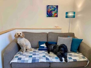 A grey sofa with a checkered blanket, two dogs sitting on it, a colorful abstract painting, and a floor lamp. - Weaver’s Den, a cosy countryside retreat (Holmfirth)