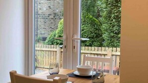 Un coin repas avec une table dressée pour deux personnes, une chaise blanche et une vue sur un mur de briques et la verdure à l’extérieur.