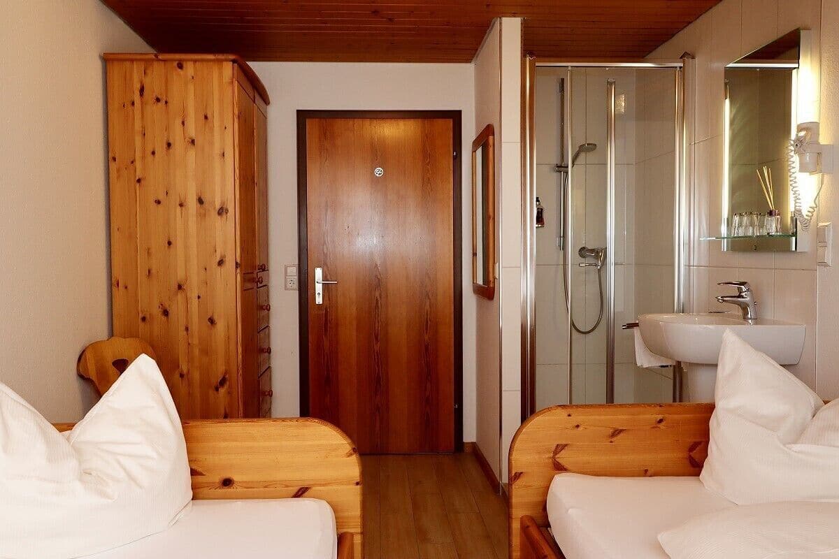 A room with two wooden beds, a wooden wardrobe, a shower area, and a bathroom with a sink and mirror.