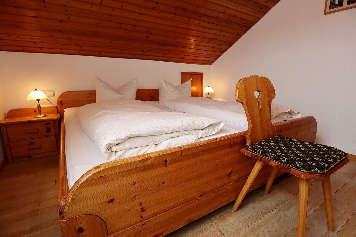 A wooden bed with white bedding, a wooden nightstand with a lamp, and a wooden chair with a patterned cushion.