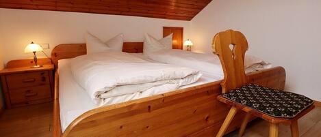 A wooden bed with white bedding, a wooden nightstand with a lamp, and a wooden chair with a patterned cushion.