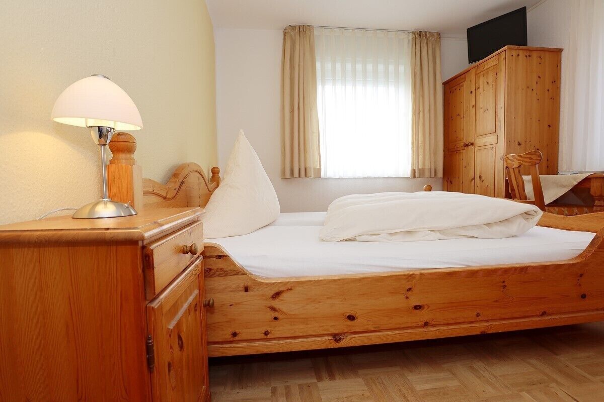 A wooden bed with a white mattress and pillows, a wooden nightstand with a lamp, a wooden wardrobe, and a window with curtains.