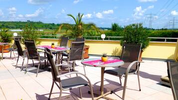 Un patio exterior con sillas y mesas de metal, rodeado de plantas en macetas y una vista despejada del campo.