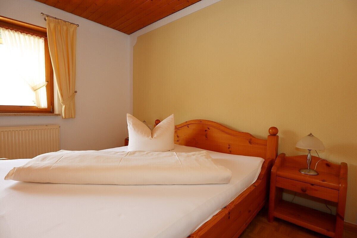 Cama bem arrumada com lençóis e almofadas brancos, cabeceira de madeira, mesa de cabeceira com abajur e janela com cortinas.