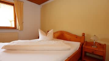 Cama bem arrumada com lençóis e almofadas brancos, cabeceira de madeira, mesa de cabeceira com abajur e janela com cortinas.