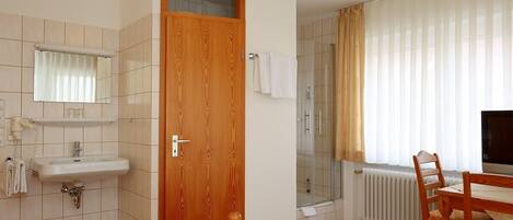 Habitación de hotel con puerta de madera, lavamanos blanco, espejo, ducha, mesa de madera con sillas y televisor.