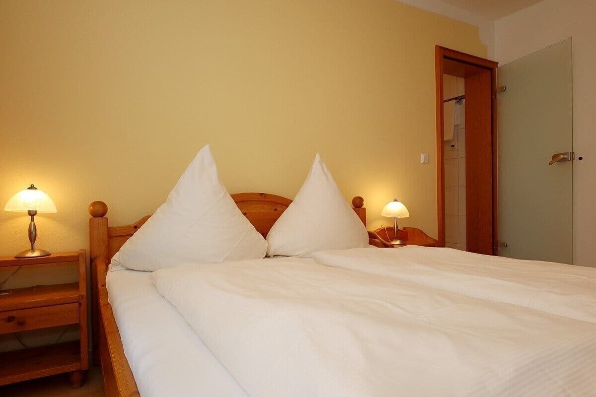 A neatly made bed with white linens and two pillows, flanked by wooden nightstands with lamps, in a room with a door and a light-colored wall.