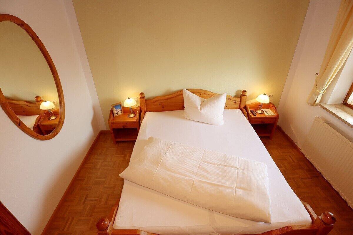 A neatly made bed with white linens, two bedside tables with lamps, a wooden headboard, a large mirror, and a window with curtains.