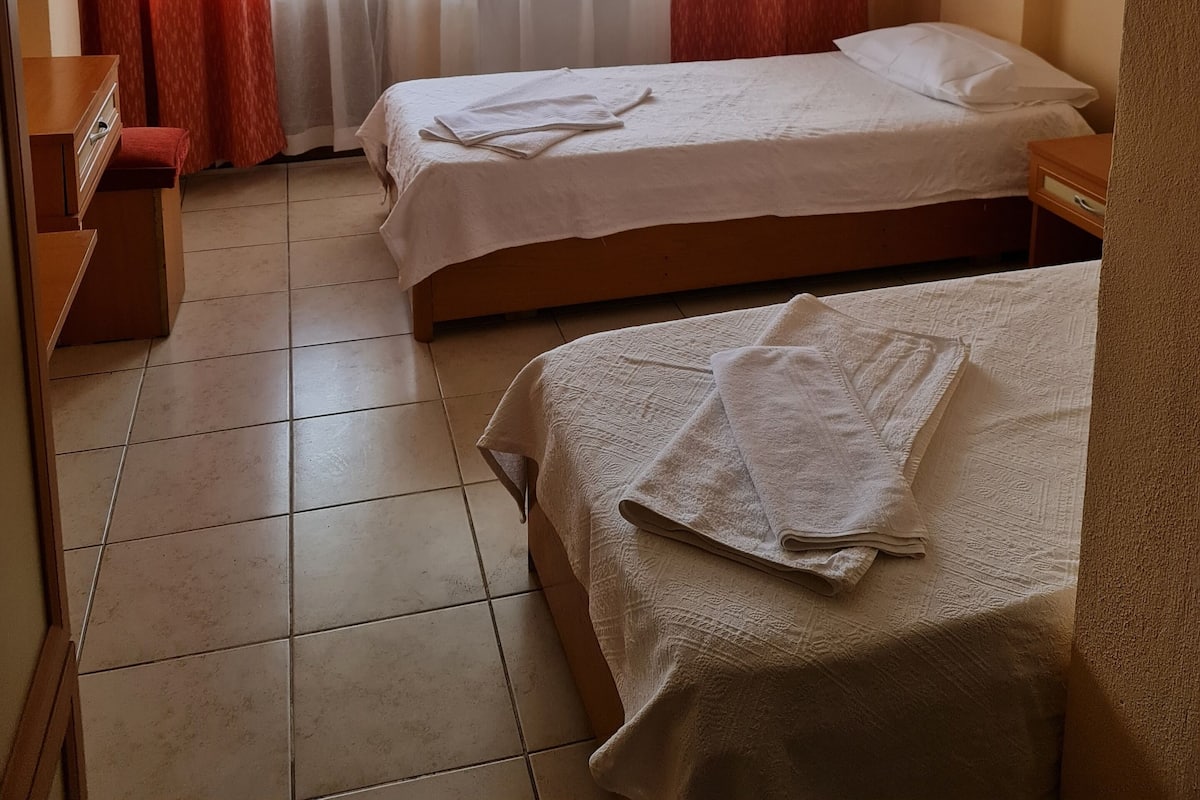 A hotel room with two beds, a wooden nightstand, a desk with drawers, and a window with sheer curtains and red drapes.