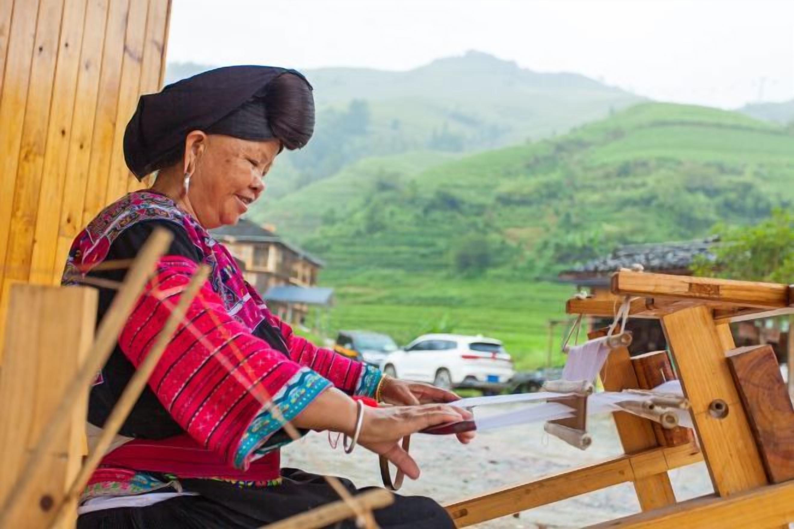 A person sitting on a wooden chair, working on a loom with a rural landscape in the background.