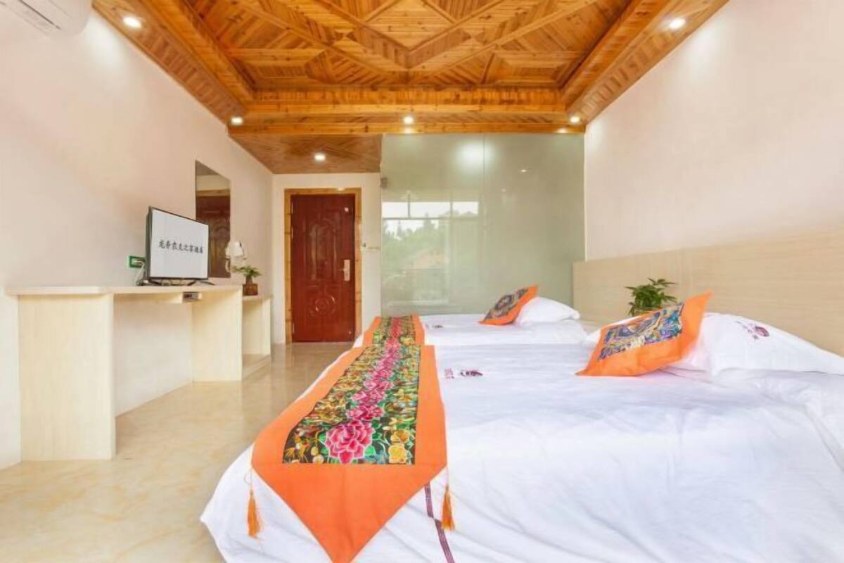 Une chambre d’hôtel avec un plafond en bois, un comptoir de réception, un lit avec une literie blanche et des oreillers orange, ainsi qu’une longue table recouverte d’un chemin de table à motifs floraux.
