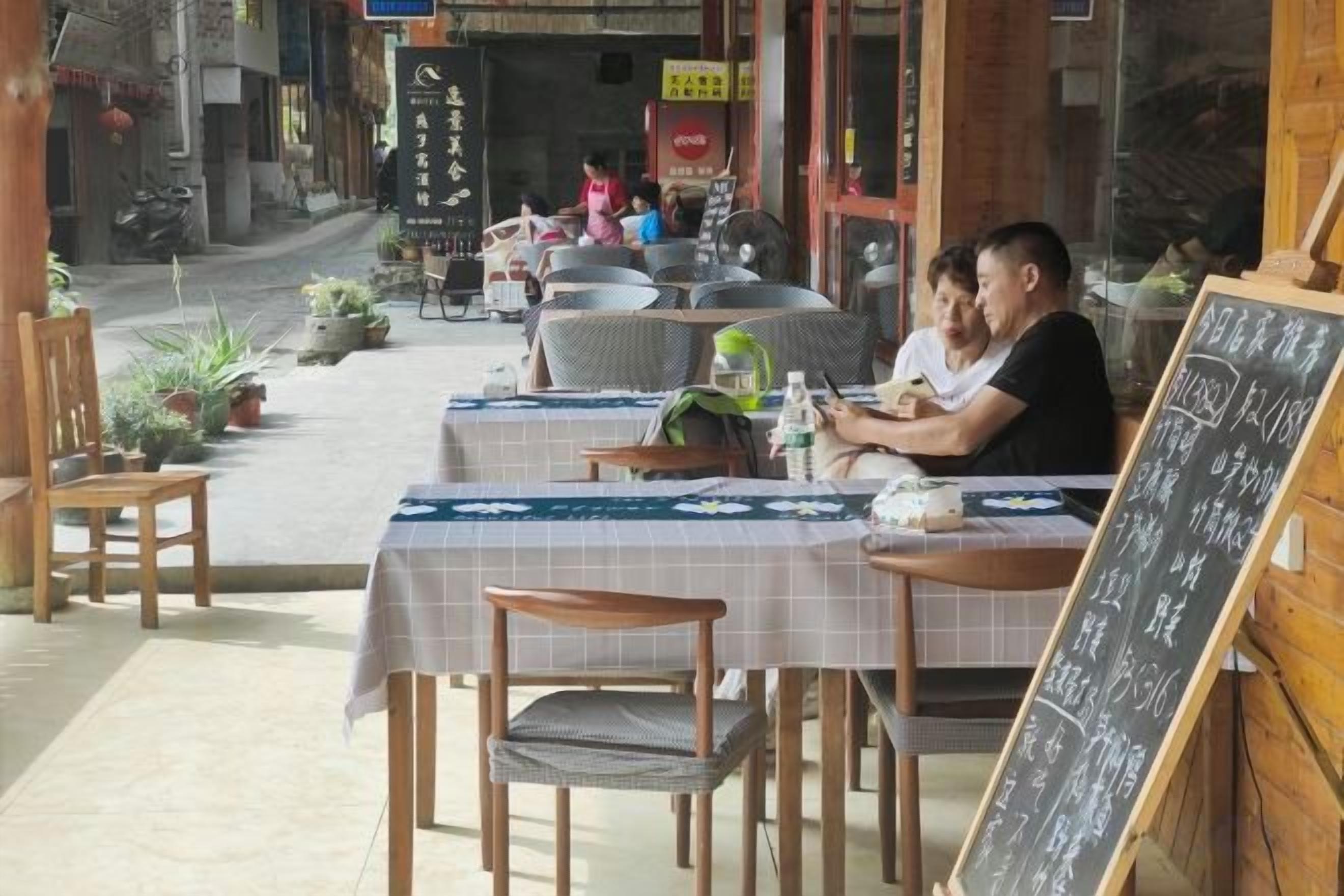 An outdoor cafe with tables and chairs, a chalkboard menu, and potted plants.