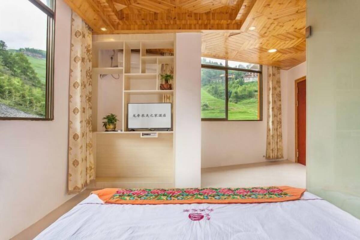 Une pièce comprenant un lit, un plafond en bois, un bureau avec des étagères et une grande fenêtre donnant sur un paysage verdoyant.