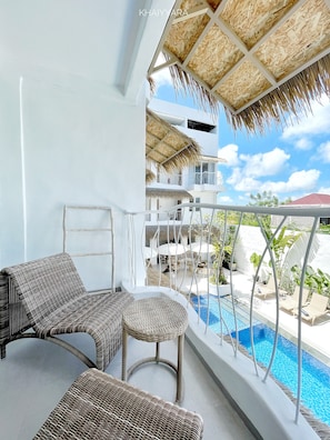 A balcony with wicker furniture, a pool, and a view of a building with a red roof. - Khaiyyara Jimbaran (Ungasan)
