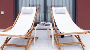 A modern building with a red facade and a sloped roof. There are two wooden lounge chairs with white cushions in front of a glass door. - Botica Guesthouse (Espinho)