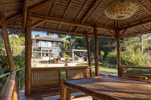 A wooden gazebo with a thatched roof, overlooking a pool area with a house in the background.