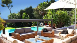 A sunlit outdoor seating area with wicker furniture, a pool, and lush greenery.