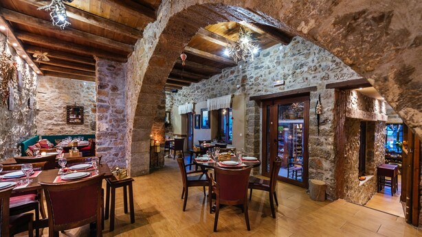 A rustic dining area with stone walls, wooden beams, and a high ceiling. Tables are set with red chairs and wine glasses. - Vasilikata (Lasithi Plateau)