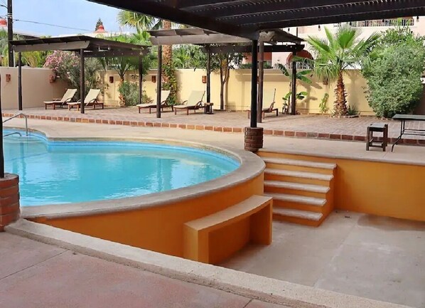 En swimmingpool med en buet kant, omgivet af en fliseterrasse og et overdækket opholdsområde med stole.