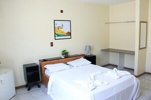 Quarto de hotel com cama, pequena escrivaninha, mesa de cabeceira, luminária, espelho e um quadro na parede.