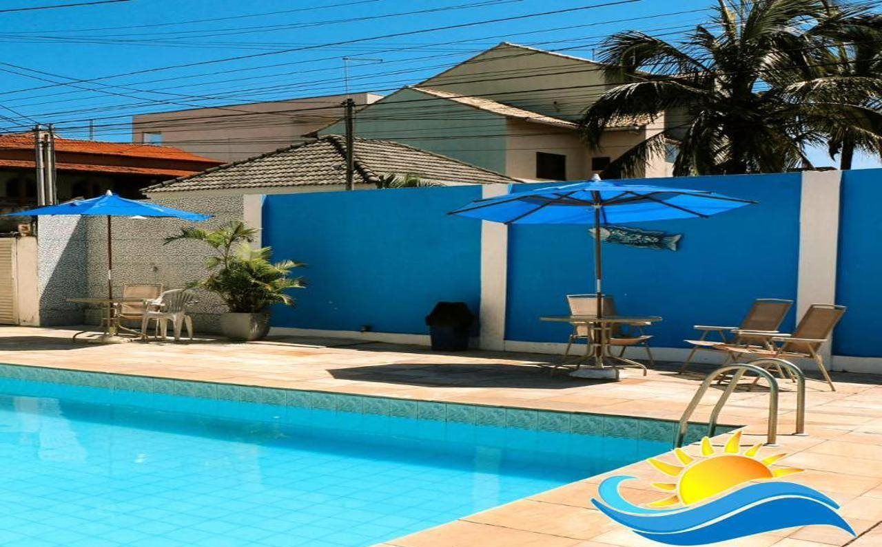 Uma piscina com guarda-sóis azuis e espreguiçadeiras, cercada por um muro azul e palmeiras.