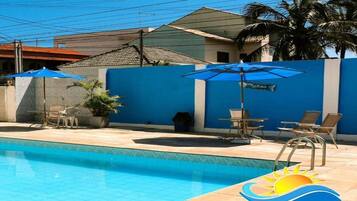 Uma piscina com guarda-sóis azuis e espreguiçadeiras, cercada por um muro azul e palmeiras.