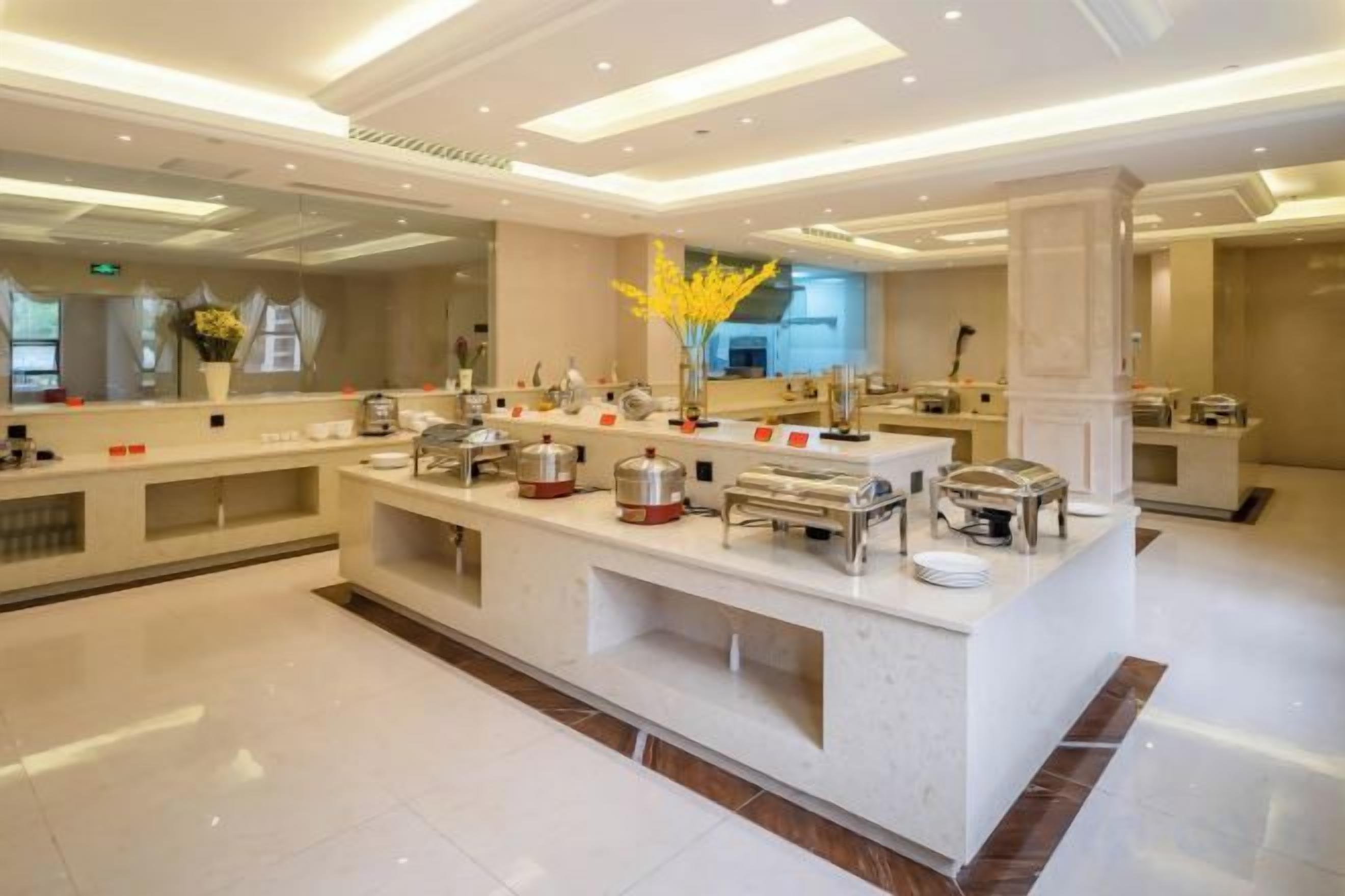 A modern hotel lobby with a central buffet area, a reception desk, and a large mirror reflecting the space.