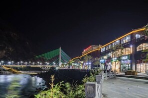 A nighttime view of a riverside area with a modern bridge, illuminated buildings, and a paved walkway. - Nujiang East Wenhao hotel (Lushui)