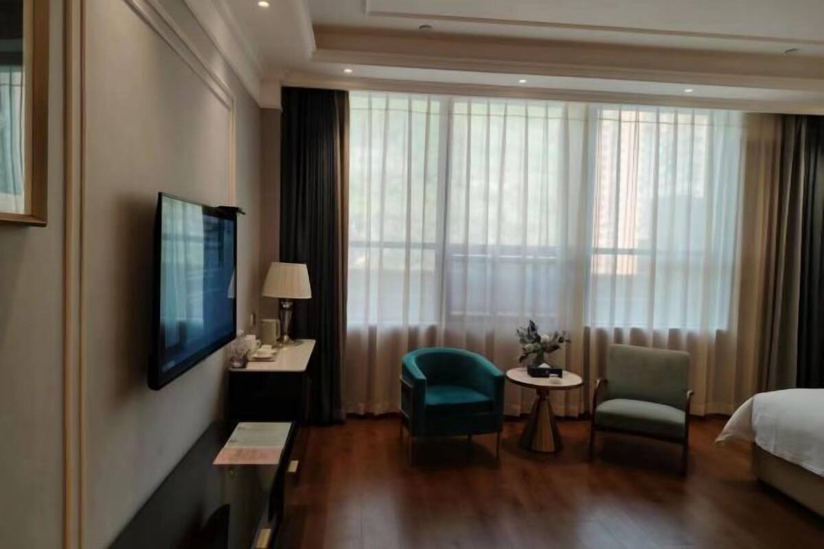 Et hotelværelse med en seng, et skrivebord med lampe, to stole, et fjernsyn og store vinduer med gardiner.