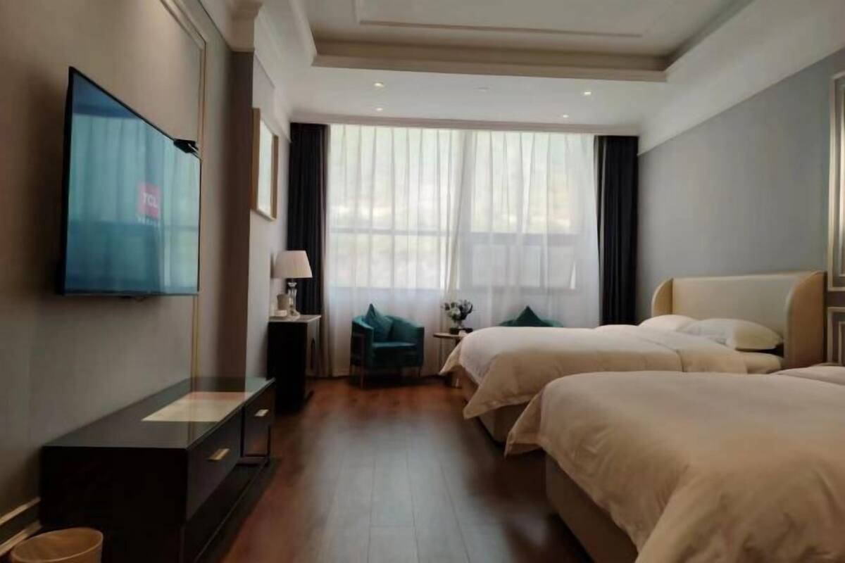 Et hotelværelse med to senge, et fjernsyn på væggen, et skrivebord med lampe samt et vindue med gardiner.